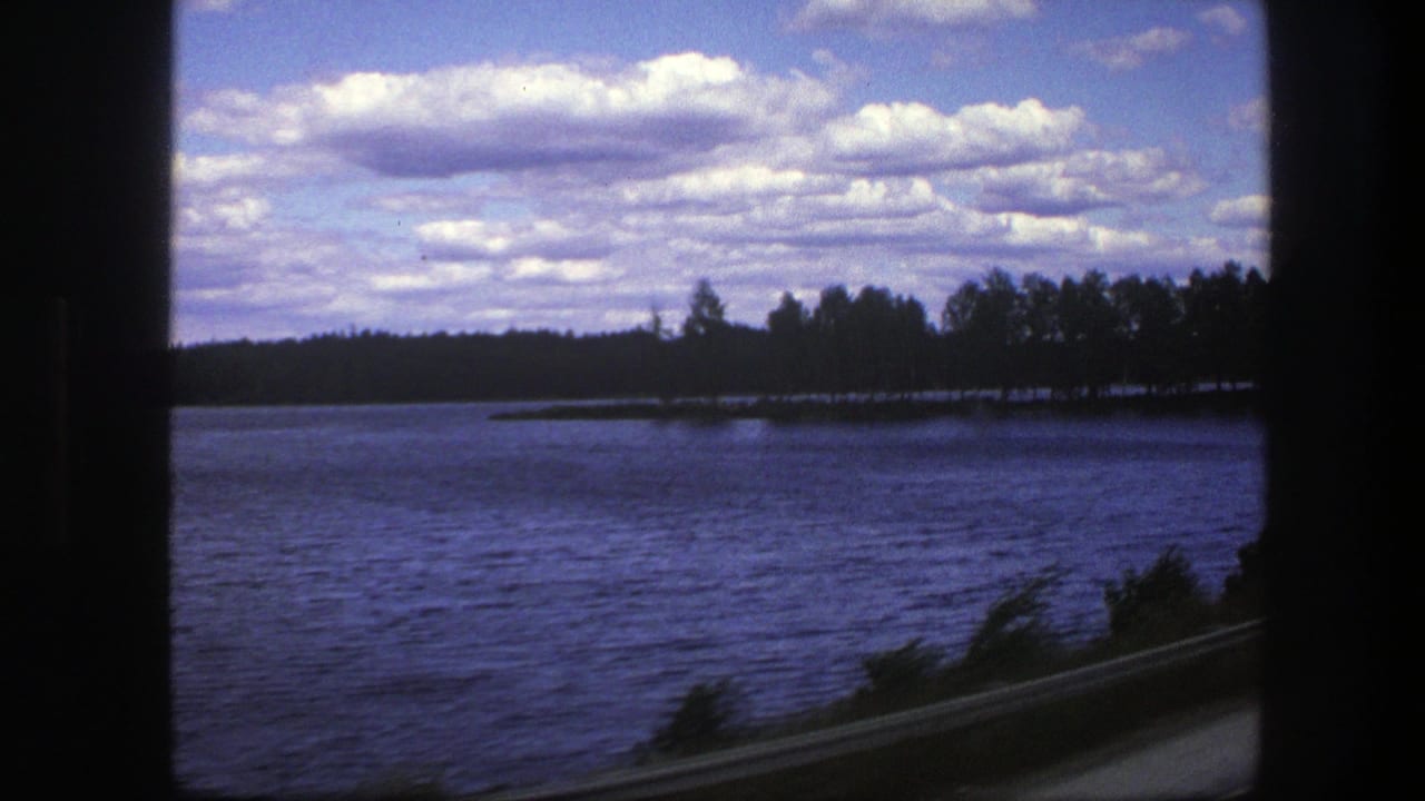 Lake beside a road with wooded shoreline and clouds overhead.