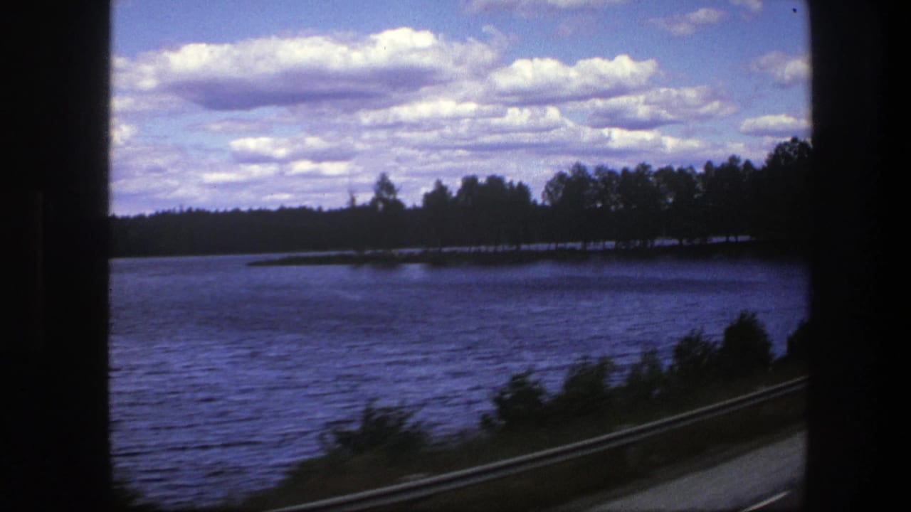 A passenger-window view in Sweden shows lake water, a low guardrail, and a tree-lined shore beneath a bright clouded sky.