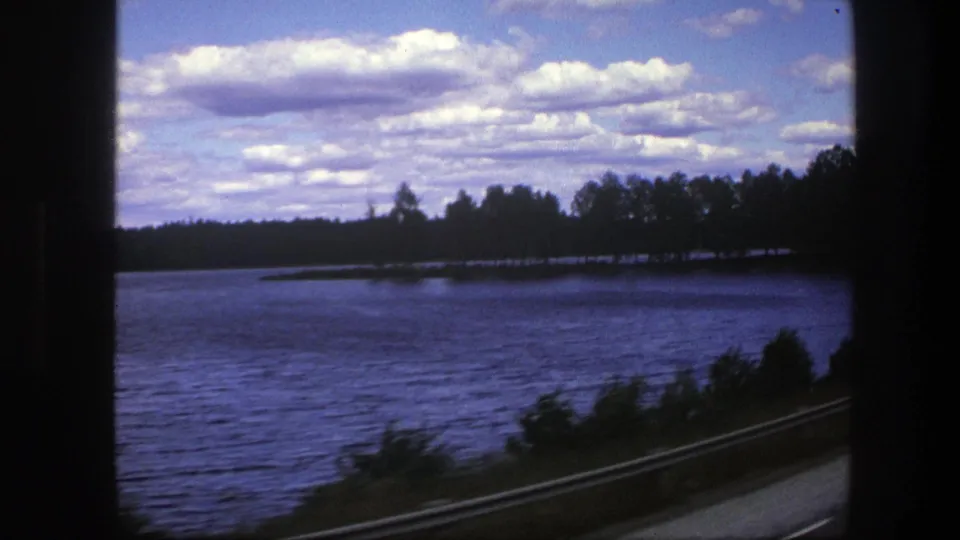 A passenger-window view in Sweden shows lake water, a low guardrail, and a tree-lined shore beneath a bright clouded sky.