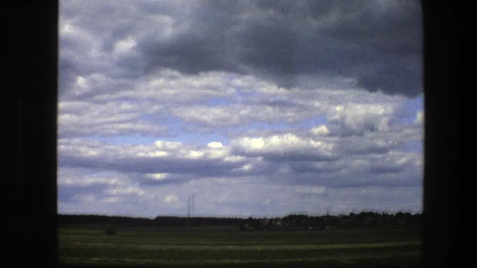 A rural Swedish field appears under layered clouds, with a low horizon and distant utility lines visible in the landscape.