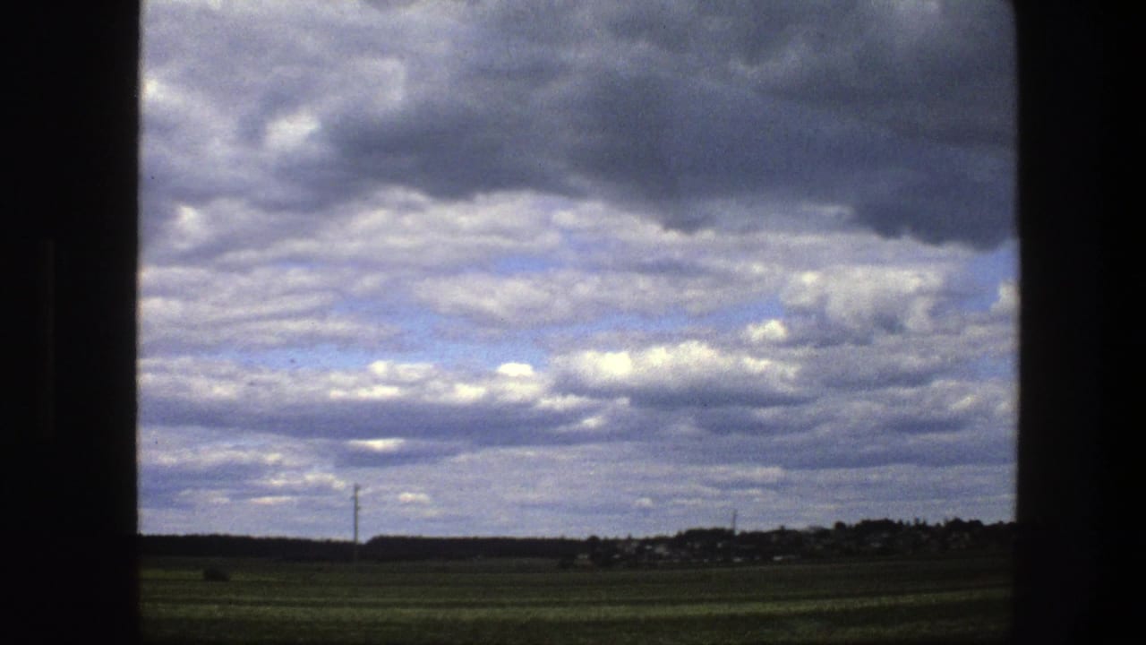 Green fields and a distant tree line seen through a dark window frame under gray and white clouds.