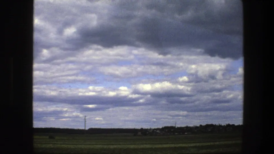1975 Swedish rural landscape seen through a vehicle window, with green fields, distant trees, small buildings, and a layered clouded sky.