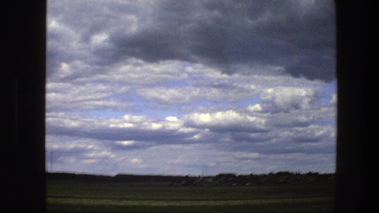 Open fields and distant buildings framed by a dark window divider beneath a cloud-filled sky.