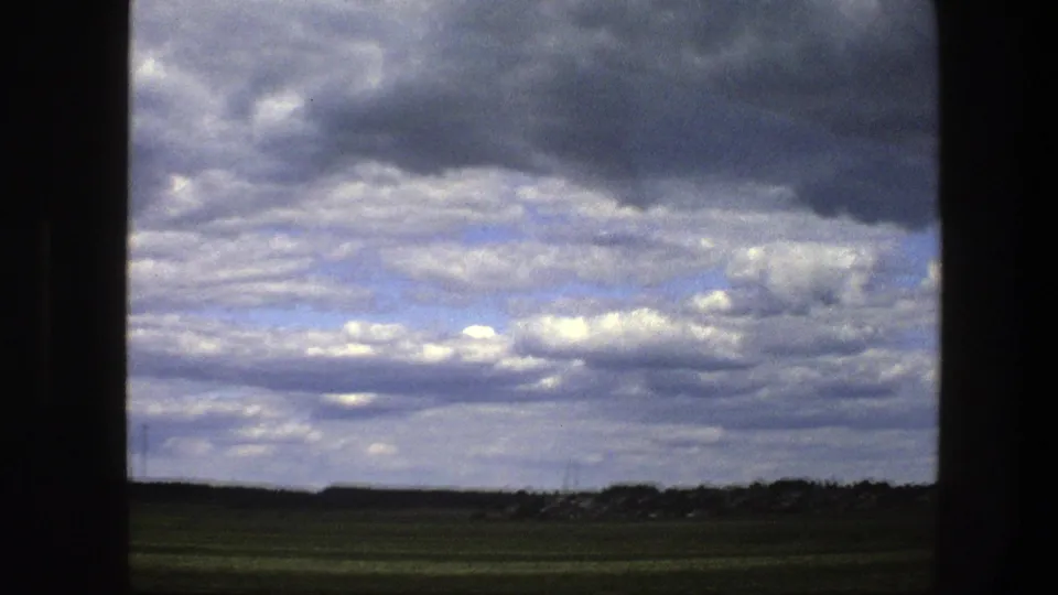 Rural Sweden in 1975 appears beyond a moving window, showing flat green fields, distant settlement edges, and white-gray clouds with blue patches.