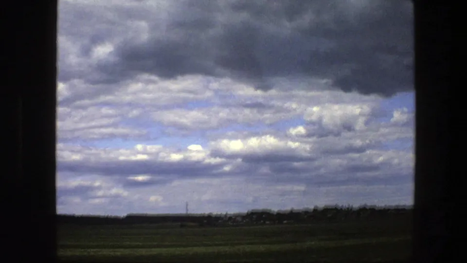 A 1975 Swedish countryside view from a moving vehicle shows green fields, a low horizon, a utility pole, and dense layered clouds overhead.