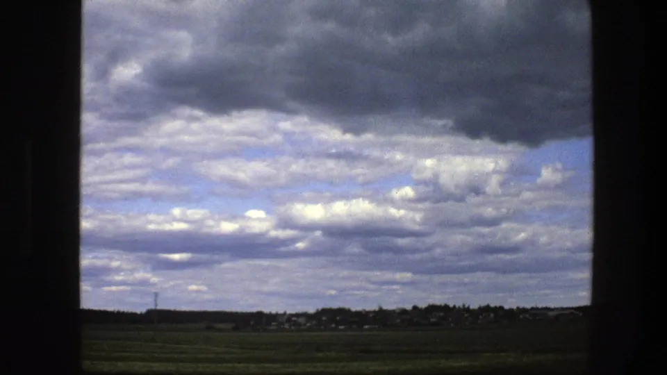 1975 Swedish rural fields pass below a clouded sky, with distant houses, a low tree line, and the dark edge of the window still visible.