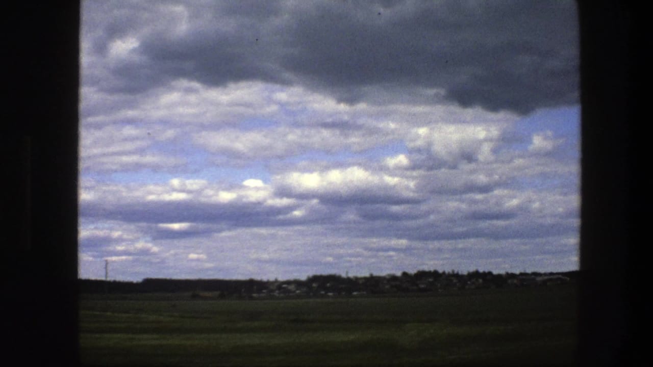 1975 Sweden home-movie frame showing open green fields, a distant settlement, and layered clouds viewed from a vehicle.