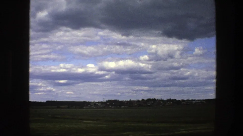 1975 Swedish countryside view from a moving vehicle, with green fields, low buildings on the horizon, and dense clouds.