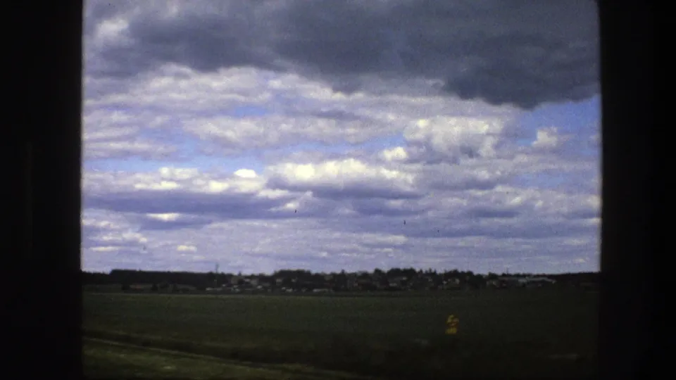 1975 Sweden travel footage frame with grassy foreground, distant settlement, treeline, and textured gray-white clouds.