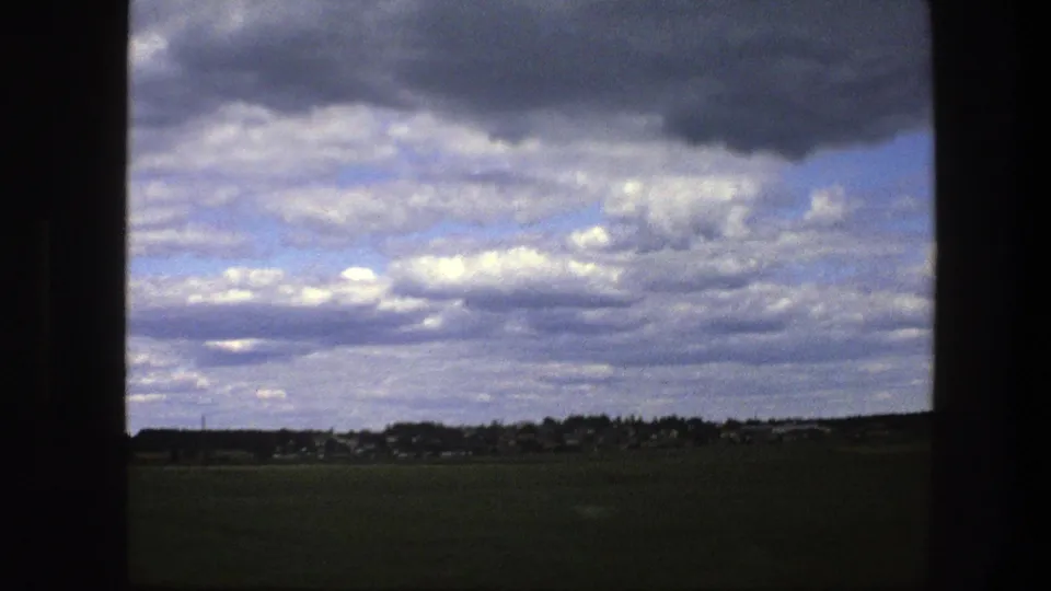 Late clip frame from 1975 Sweden showing a vehicle-framed rural field, low settlement, and expansive cloudy sky.