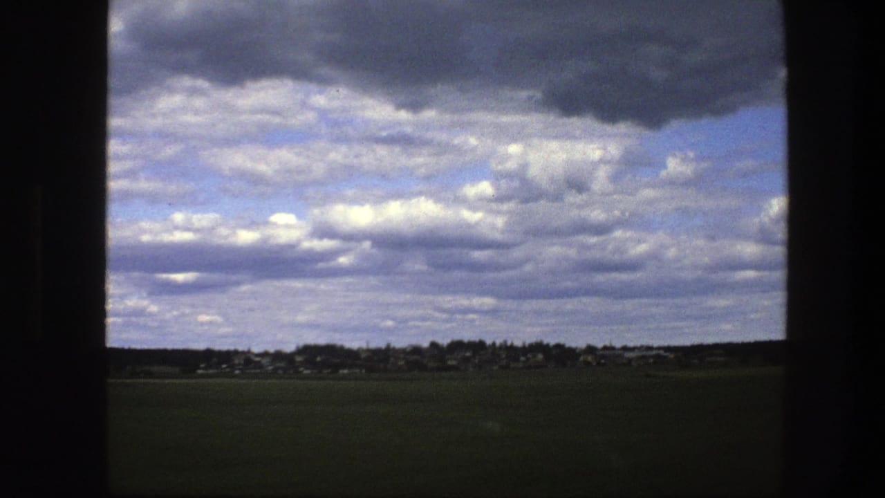 Green field and distant village beneath a cloudy sky, partly framed by dark vertical edges.