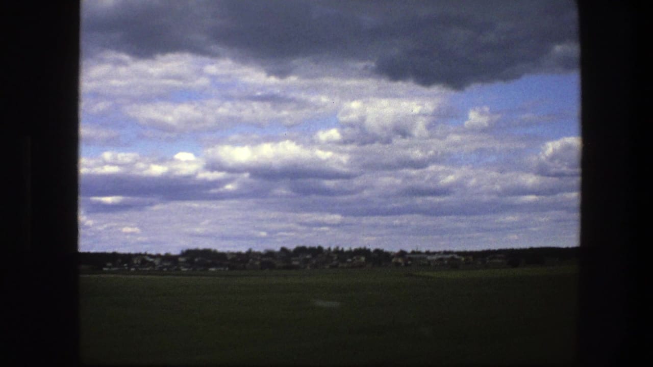 Cloud-filled sky over a dark green field and distant settlement, seen through a vertical frame.