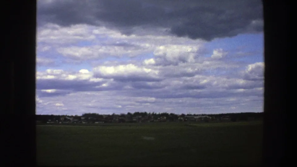 Shaky 8mm view in Sweden, 1975, showing a green field, distant village line, and textured grey-white clouds.