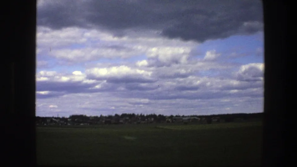 1975 Swedish countryside view with a low village horizon and dramatic cloud cover, framed by dark window or structural edges.