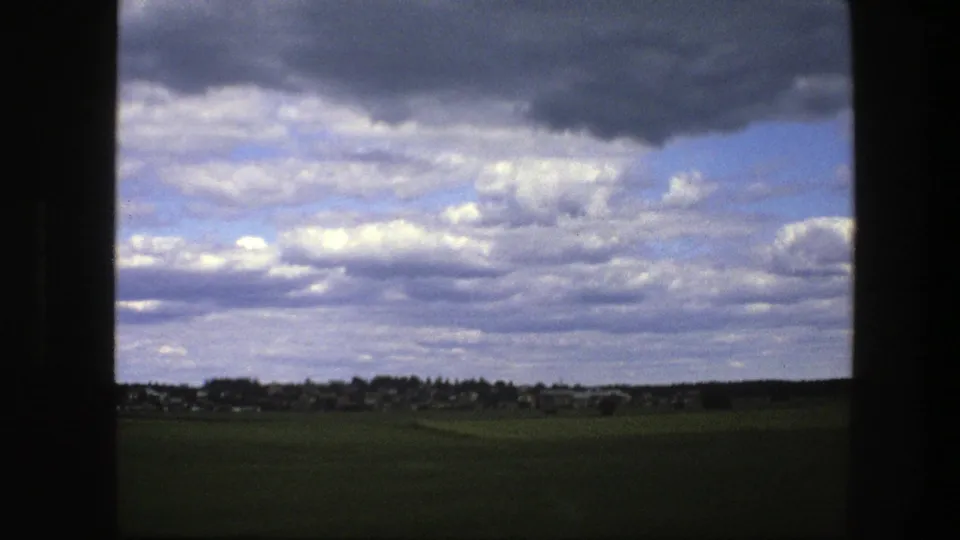 1975 Swedish rural field and distant village under heavy clouds, with a small dark moving blur near the lower right foreground.