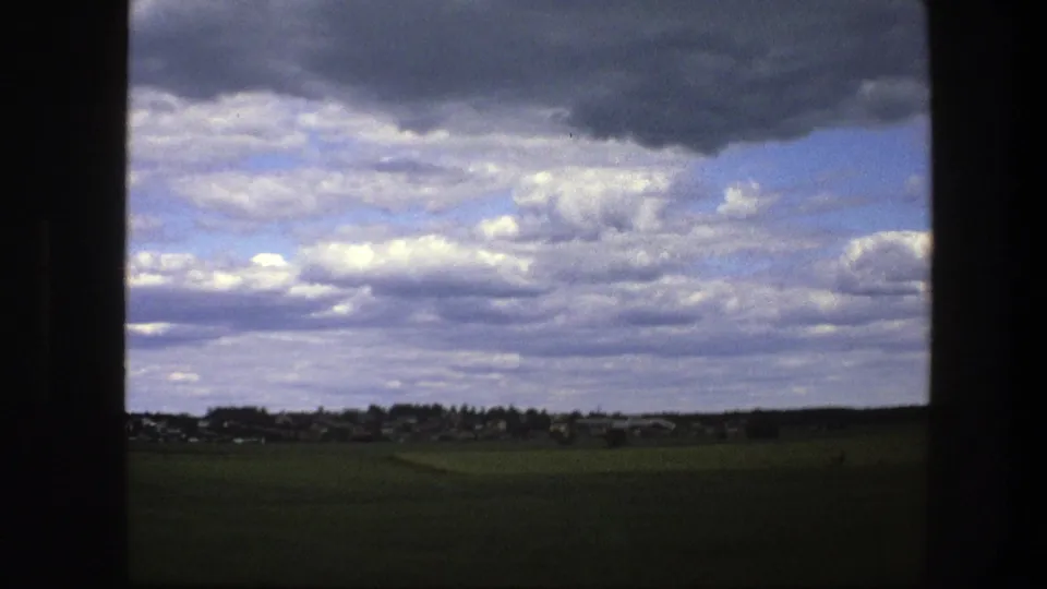 1975 Swedish home-movie view of green fields and a distant settlement under layered clouds, filmed from a moving vantage point.