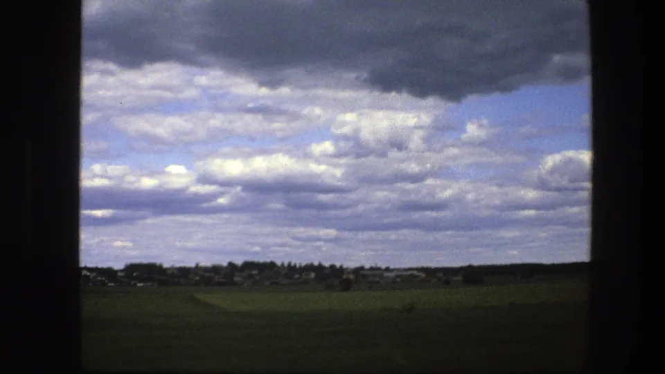 Swedish rural landscape with open fields, low buildings on the horizon, and textured cloud cover in 1975 8mm footage.