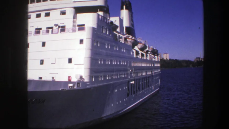 Large white cruise or passenger liner seen close to the hull on blue water, with rows of windows and deck equipment visible.