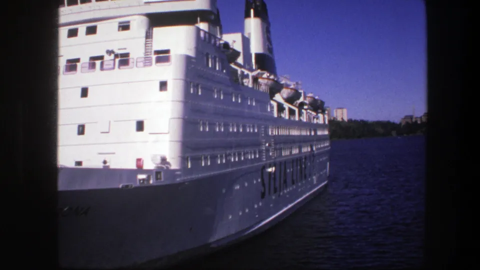 1975 Sweden 8mm frame showing a large white passenger ship on a river, with lifeboats, deck windows, distant trees, and buildings.