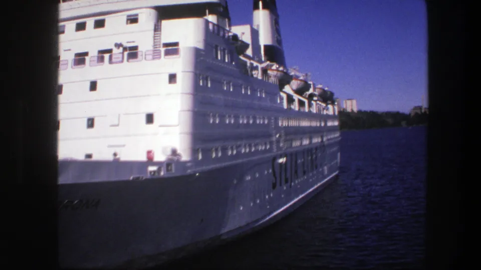 Close 1975 Sweden 8mm view of a white passenger ship, with deck windows, lifeboats, and shoreline visible beyond the river.