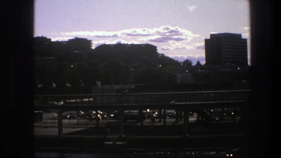 1975 Sweden 8mm frame looking across an urban waterfront, with bridge traffic, city buildings, and distant hills beyond the port area.