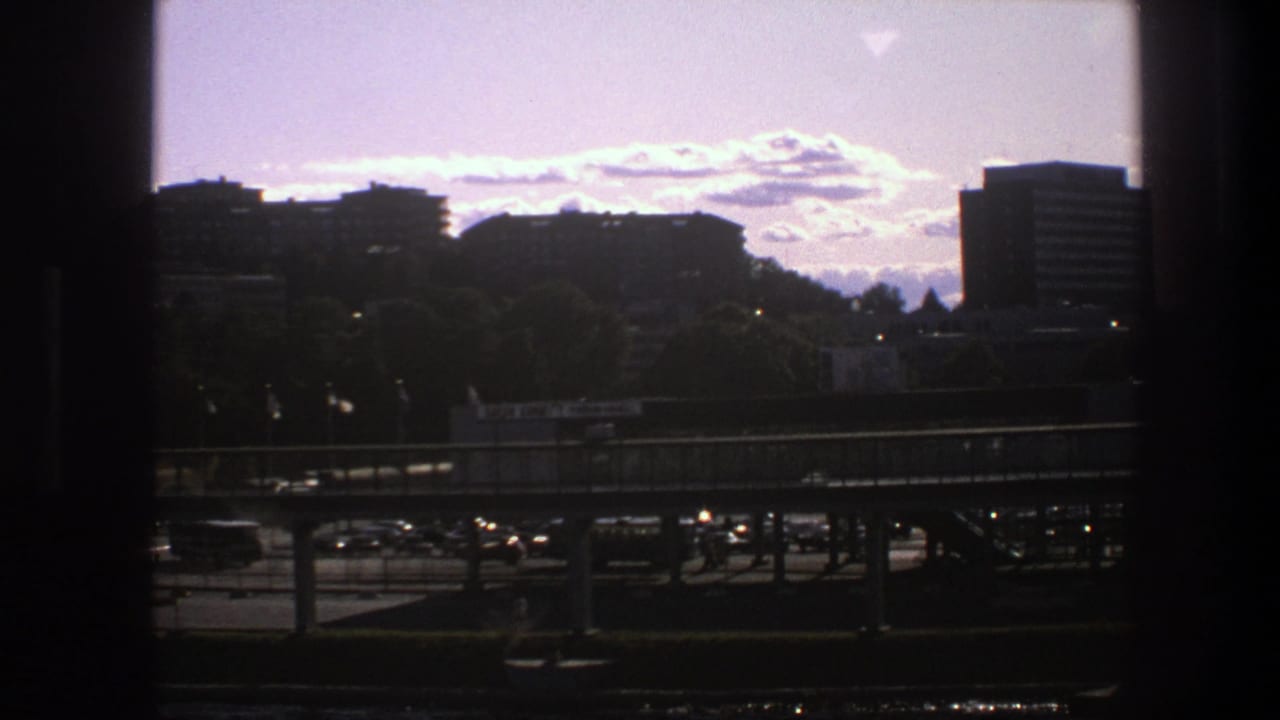 1975 Sweden 8mm frame showing a dark window edge around distant city buildings, trees, an elevated road, and light traffic under a bright sky.