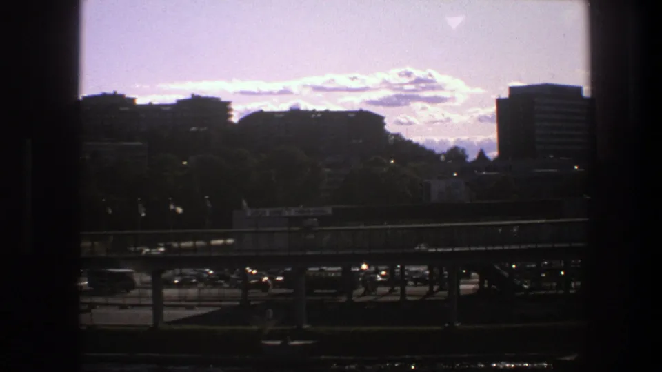 1975 Sweden 8mm frame showing a dark window edge around distant city buildings, trees, an elevated road, and light traffic under a bright sky.