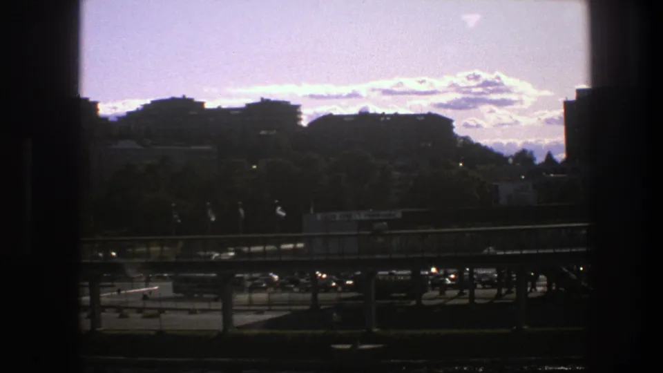 1975 Sweden urban view with silhouetted window framing, low buildings, trees, an elevated roadway, and small vehicles in sunlight.