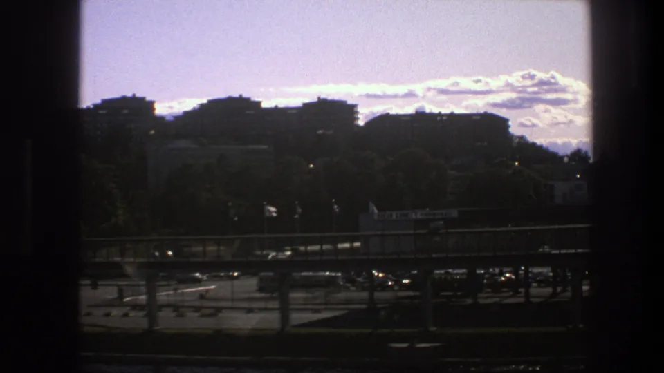 1975 Sweden 8mm view of shadowed urban buildings, roadside trees, an elevated road, and scattered cars beneath a bright clouded sky.
