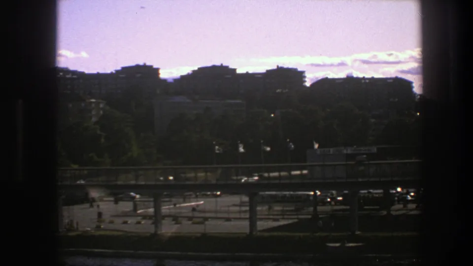 1975 Sweden city frame with dark vertical borders, distant buildings, trees, elevated roadway, and small vehicles on sunlit streets.