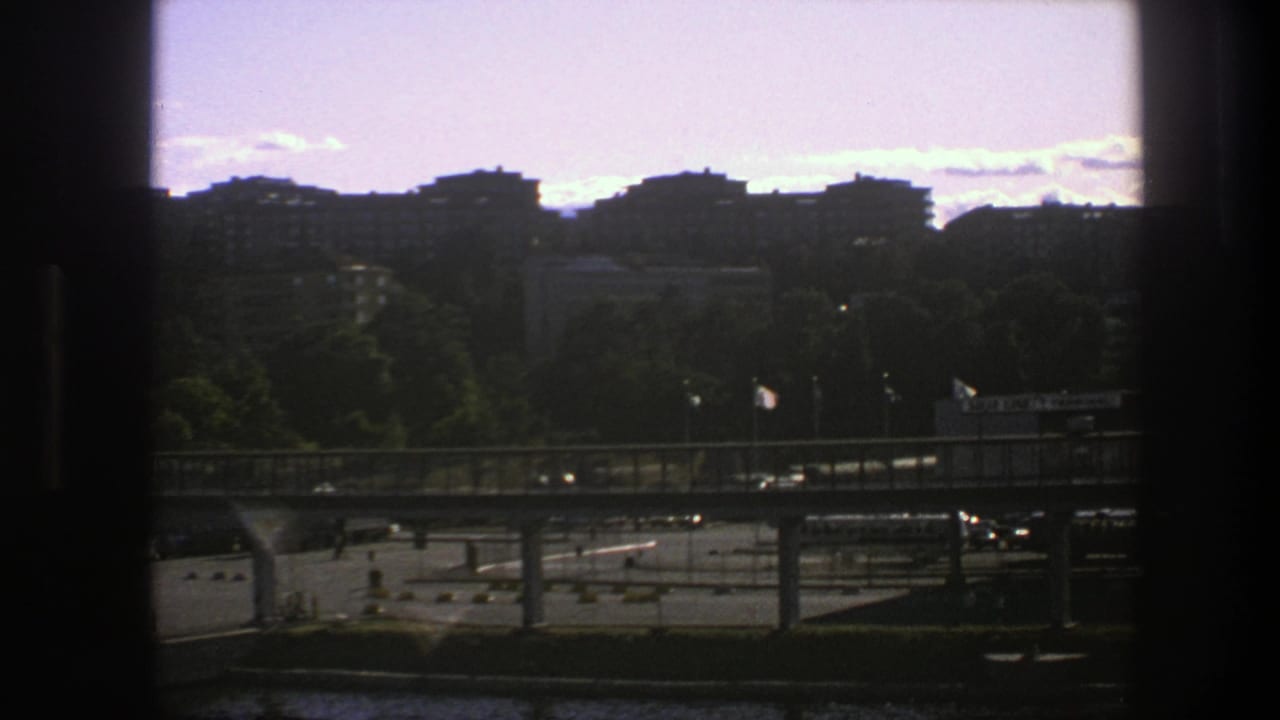 City bridge and waterfront seen through a dark window frame, with apartment blocks above trees.