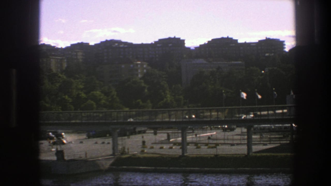 1975 Sweden city view with a bridge, moving cars, flags, treeline, and apartment blocks beyond the waterfront.