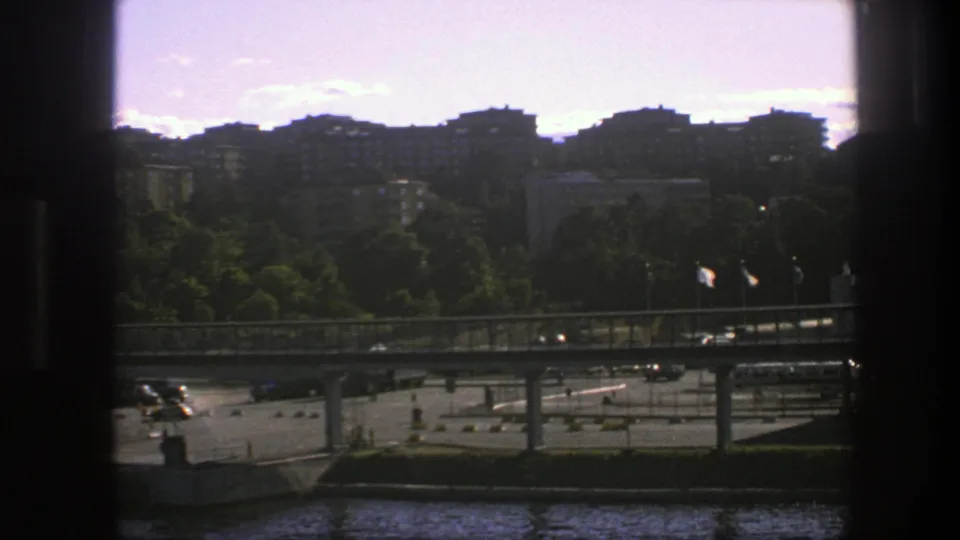 1975 Sweden city view with a bridge, moving cars, flags, treeline, and apartment blocks beyond the waterfront.