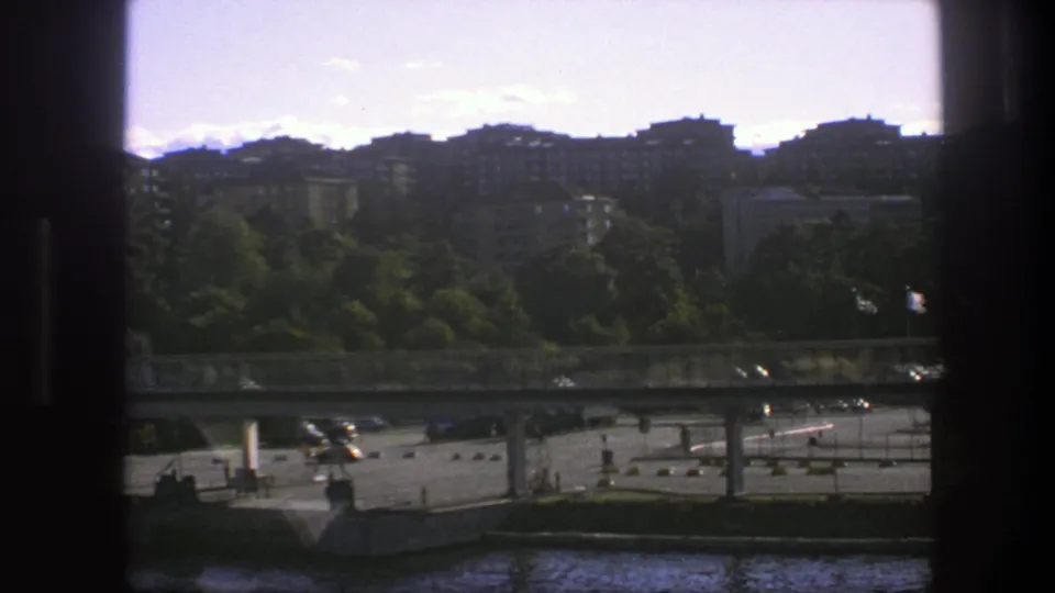1975 Sweden 8mm view across water toward a bridge, paved parking or traffic area, trees, and distant residential blocks.