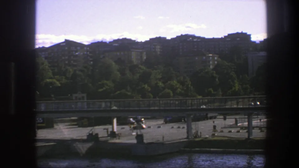 1975 Sweden home-movie view from a moving boat, showing a bridge, waterfront traffic, trees and tan apartment buildings.