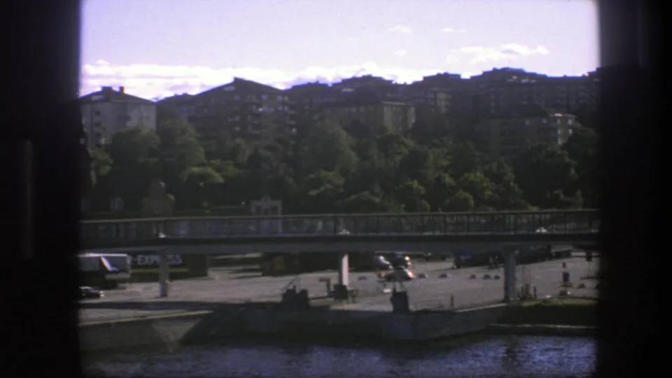 Moving 8mm view of a Swedish waterfront with a concrete bridge, green treeline, apartment blocks and passing road traffic.