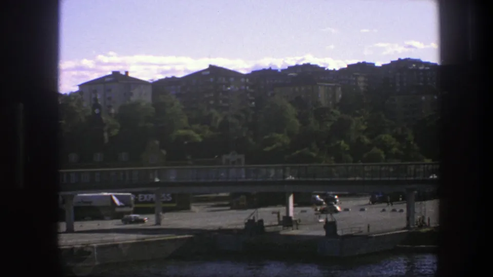 1975 Sweden waterfront scene with a white lettered van, bridge supports, road traffic, trees and apartment buildings beyond.
