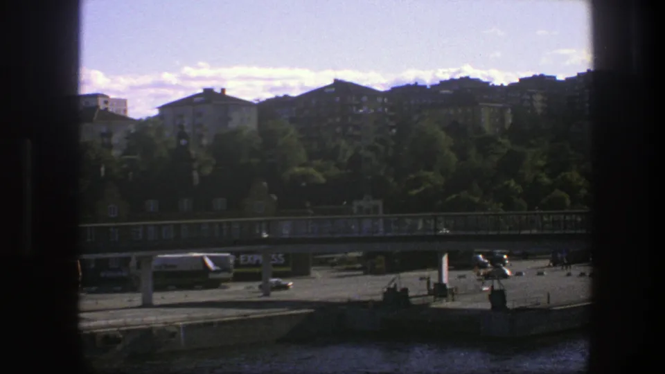 Swedish 1975 boat-view cityscape with water in the foreground, a bridge and road below trees and tan apartment buildings.