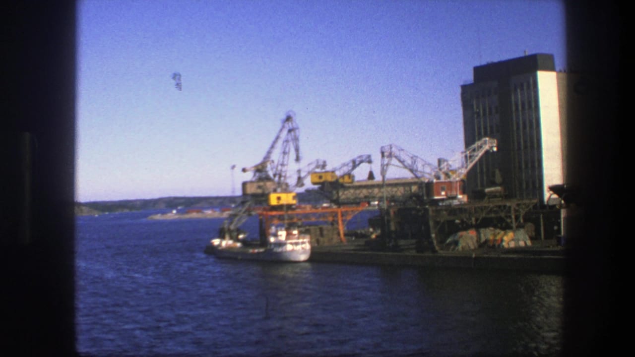 1975 Sweden 8mm harbor view showing cranes beside a moored vessel, open blue water, distant shore, and a tall industrial building.
