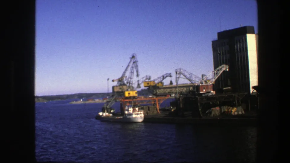 1975 Sweden 8mm port scene with multiple cranes, dock machinery, a vessel at the quay, blue bay water, and an industrial building.