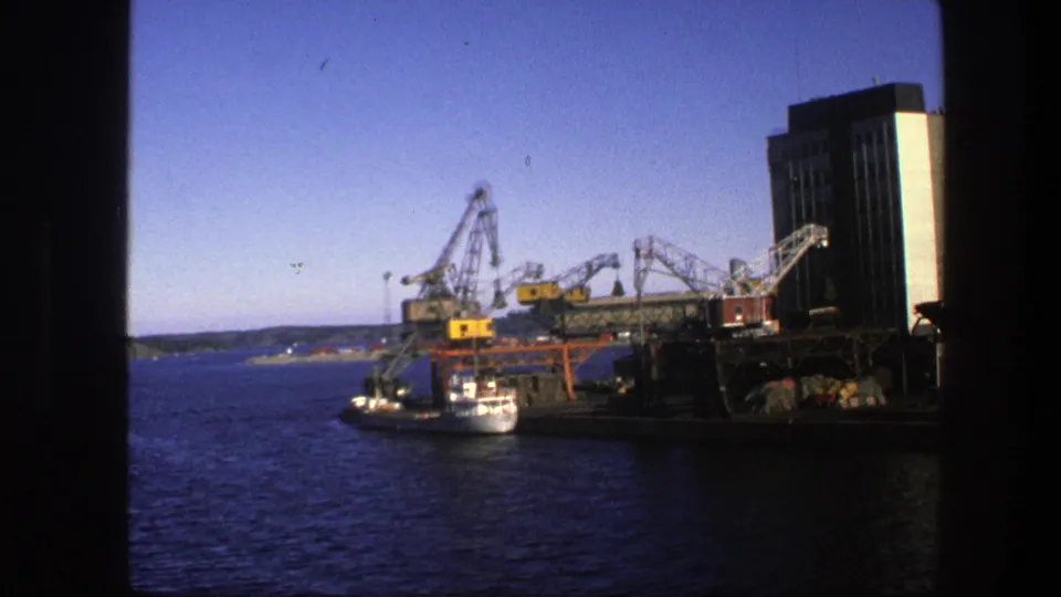 1975 Sweden 8mm view of an industrial harbor with cranes over a docked vessel, open water, distant land, and a tall port building.