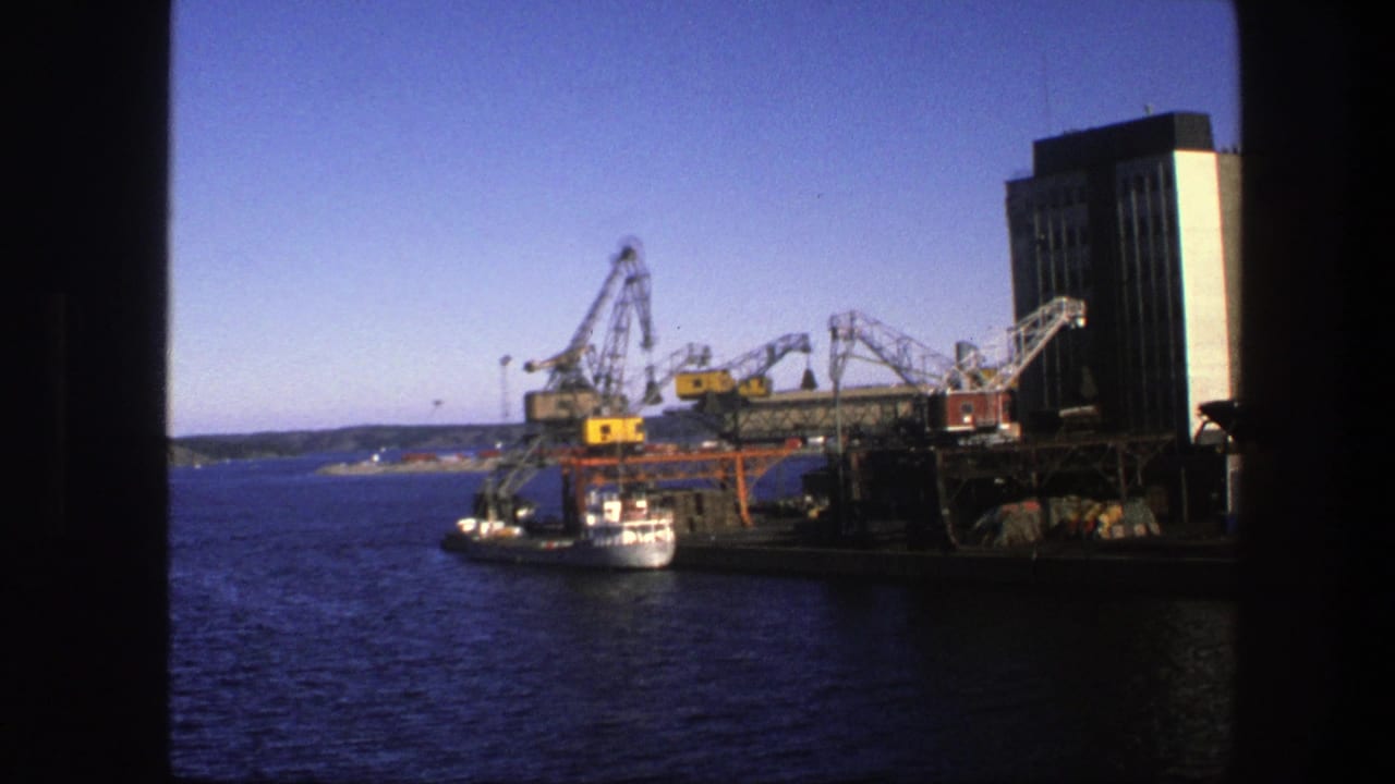 Industrial waterfront with cranes, a tall building, a small boat, and blue water framed by dark edges.