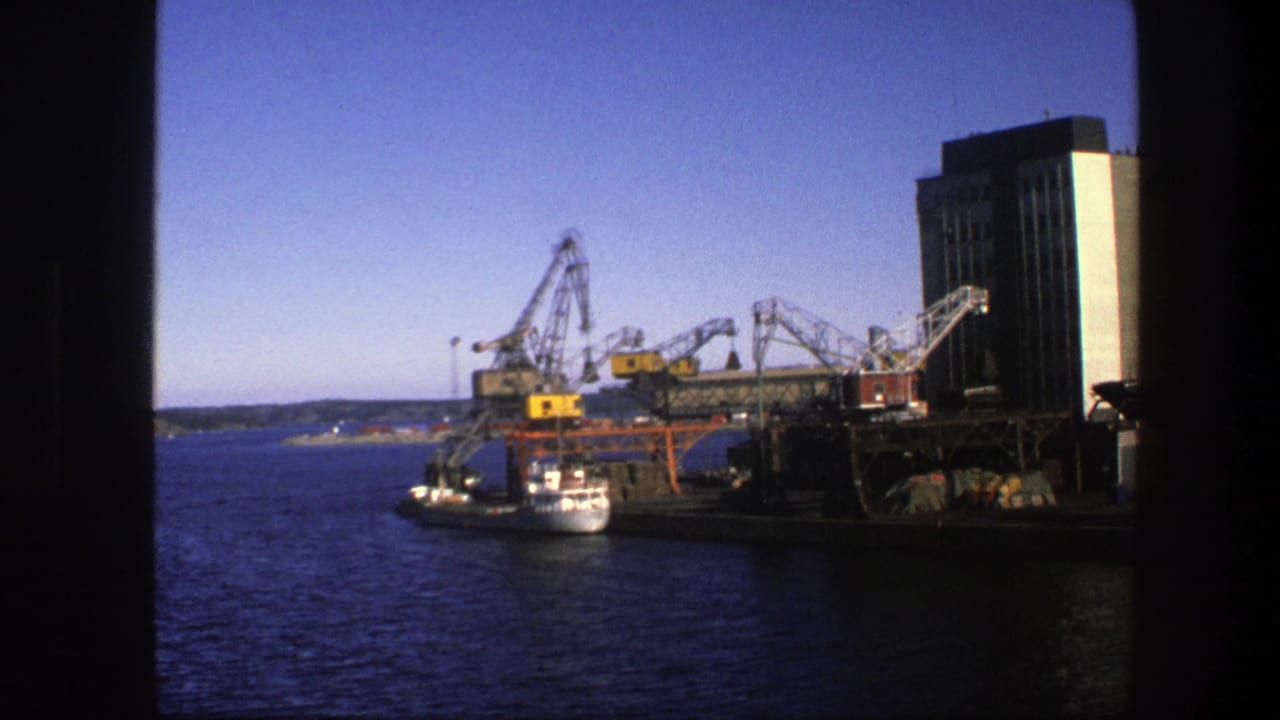 1975 Sweden home-movie frame of an industrial dock with cranes and a tall building, seen across water through dark framing.