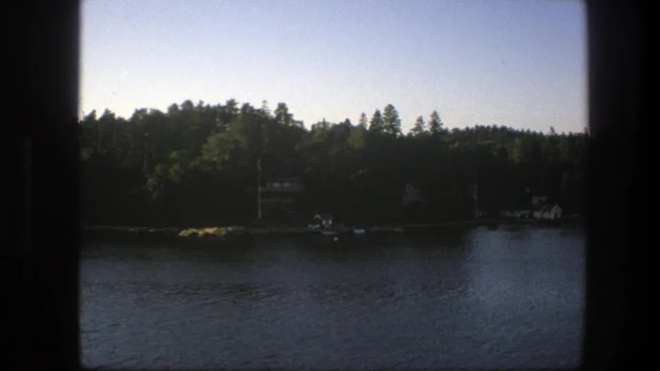 1975 Sweden 8mm frame showing a quiet wooded lakeshore with water, small houses, and utility poles in subdued exposure.