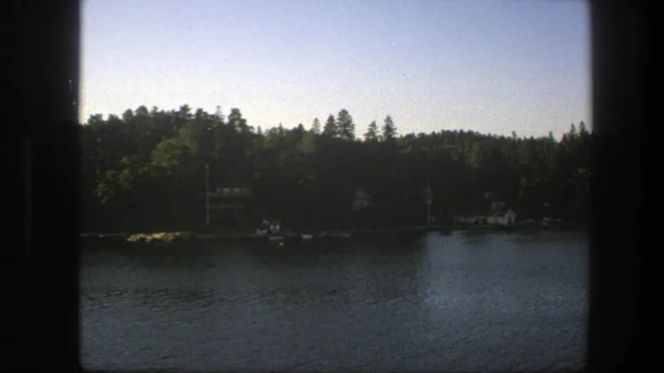 1975 Sweden home-movie frame of calm water and a forested lakeshore with distant houses, seen through dark edge framing.