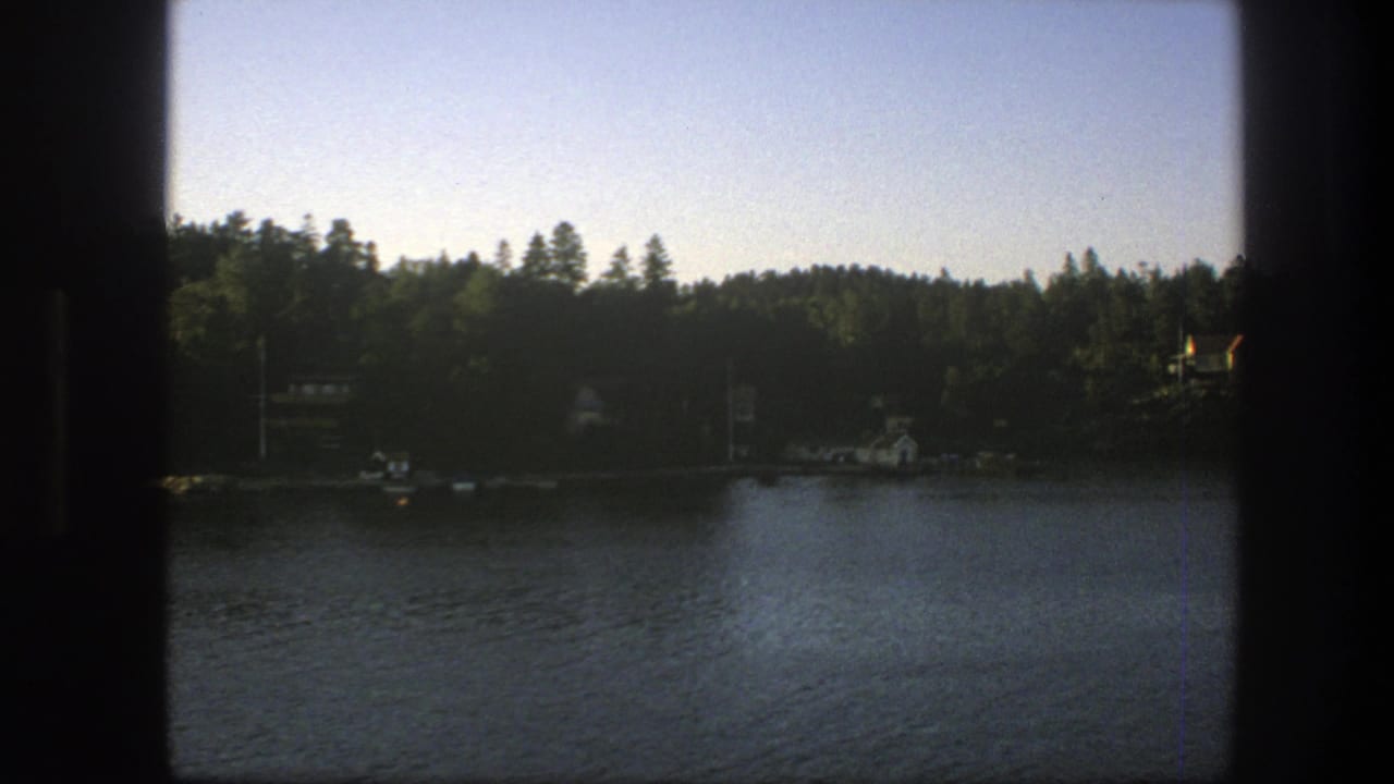 Swedish lake shoreline in 1975, filmed through a dark boat-window edge with trees, water, and distant houses visible.