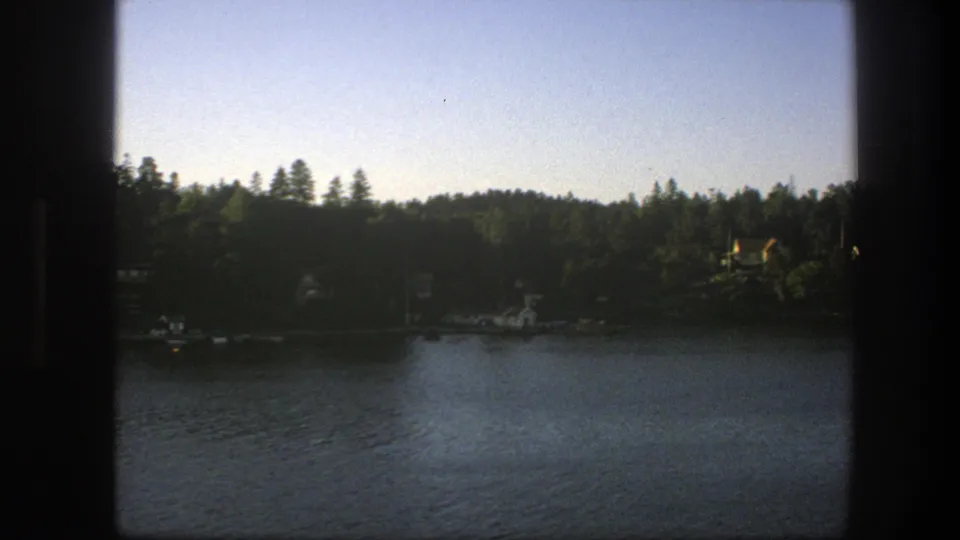 A 1975 Swedish lake scene from a moving boat, with a tree-covered bank, small shore buildings, and soft late-day light.