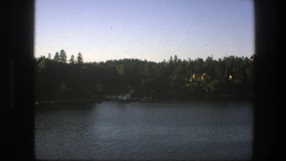 Sweden 1975 home-movie still showing a broad lake foreground and wooded shoreline under a pale late-afternoon sky.