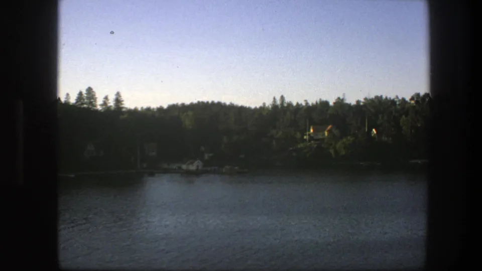 1975 8mm Sweden footage showing water in the foreground, dense trees across the shore, and small houses partly visible through the frame trim.