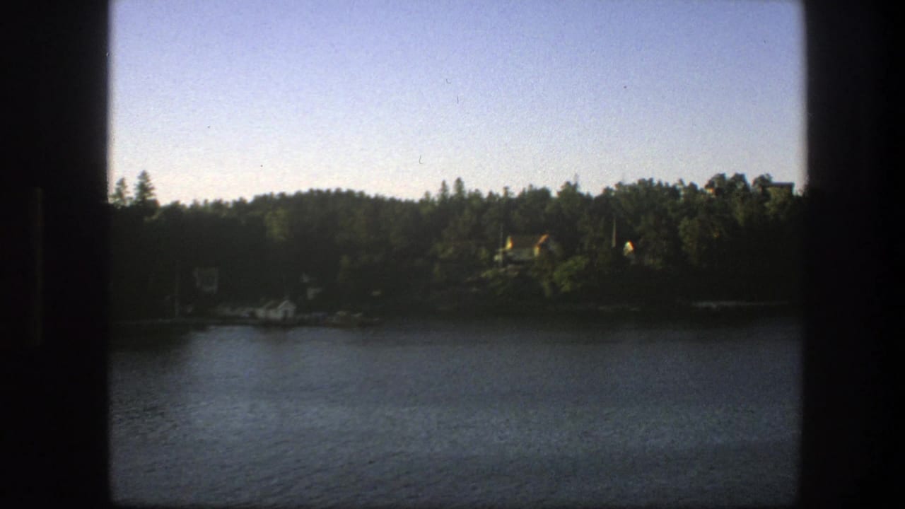 A Swedish lakeside shore appears beyond rippled water, with small houses tucked among trees and dark boat-window edges framing the scene.